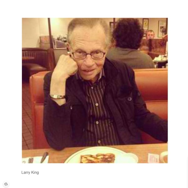 Larry King in glasses, wearing a striped shirt and jacket, sitting in a red booth at Canter's Deli.