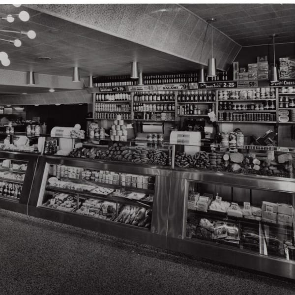 Black and white photo of a deli counter with various meats, cheeses, and canned goods on display.