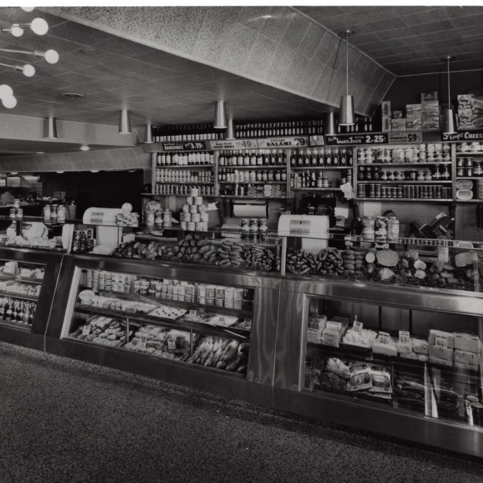 Black and white photo of a deli counter with various meats, cheeses, and canned goods on display.