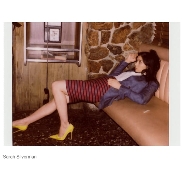 Sarah Silverman reclines on a diner booth next to a payphone, wearing a striped skirt and yellow heels.