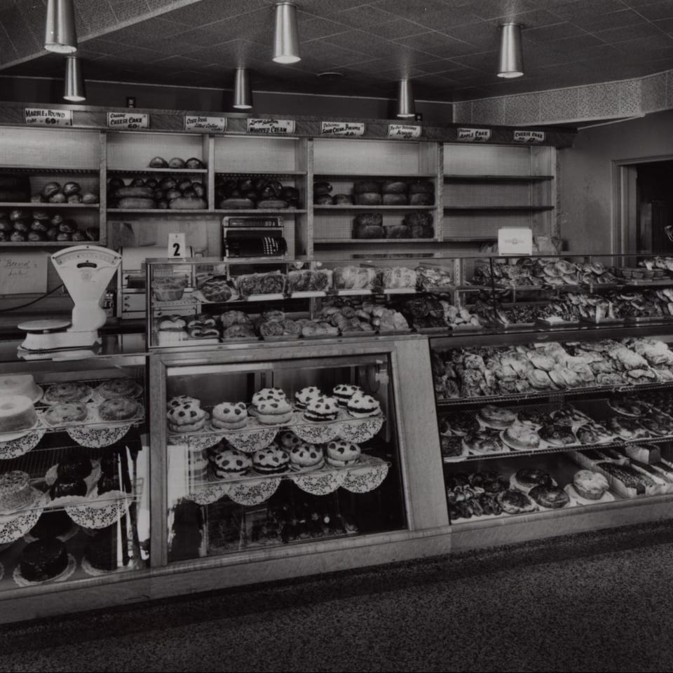 Black and white photo of a deli bakery display case filled with various breads, cakes, and pastries.