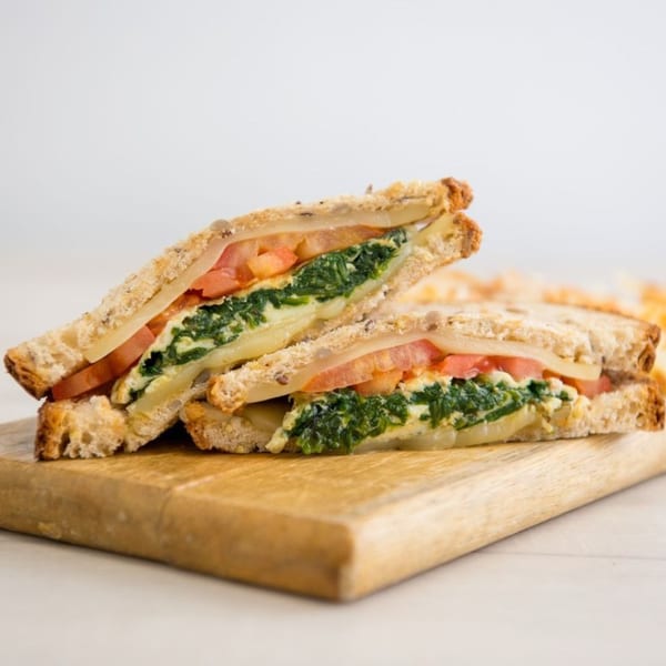Two halves of a grilled cheese sandwich with spinach and tomato on a wooden board.