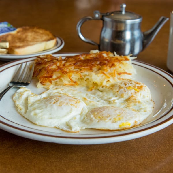 A deli breakfast of fried eggs, hash browns, and toast with butter, next to a metal teapot.